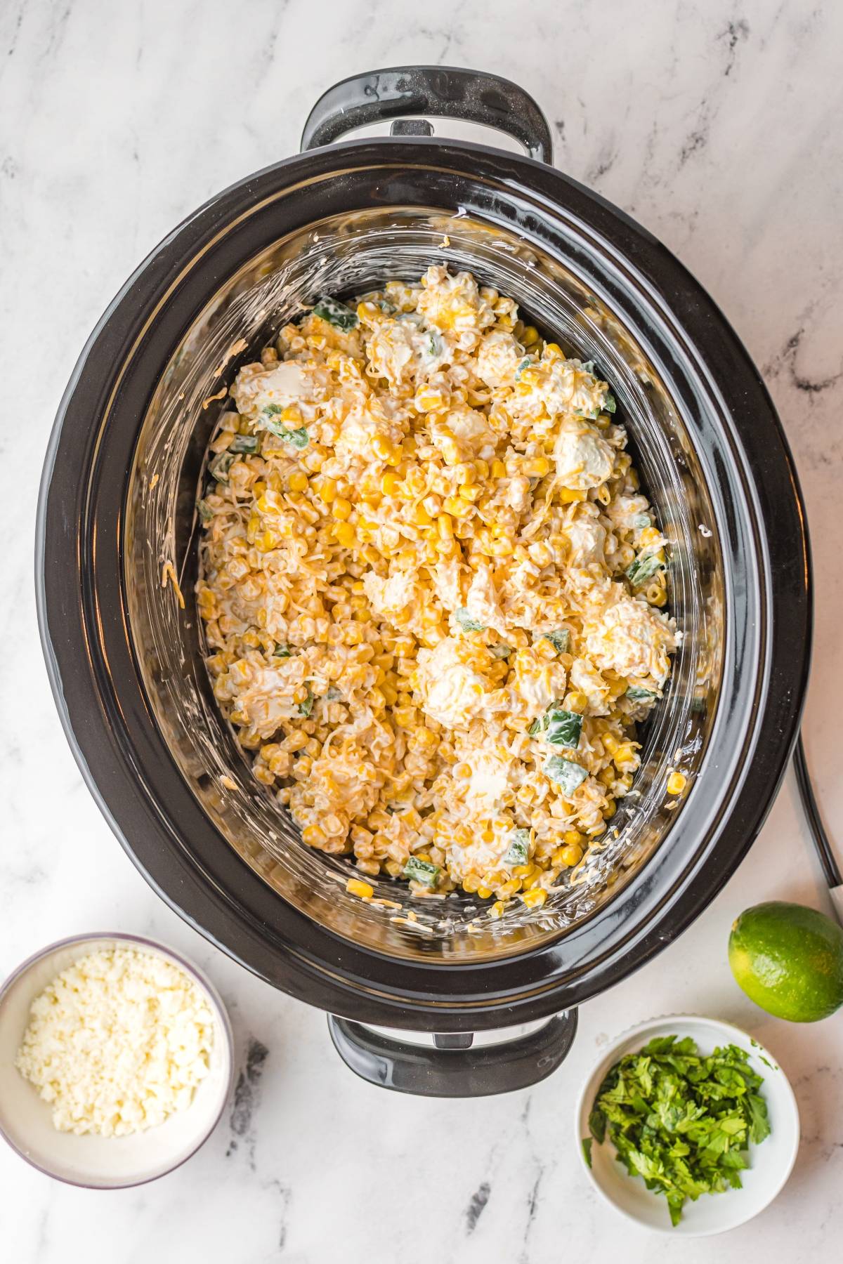 Slow cooker filled with creamy easy street corn dip, with bowls of cheese and chopped cilantro beside it on a marble surface.