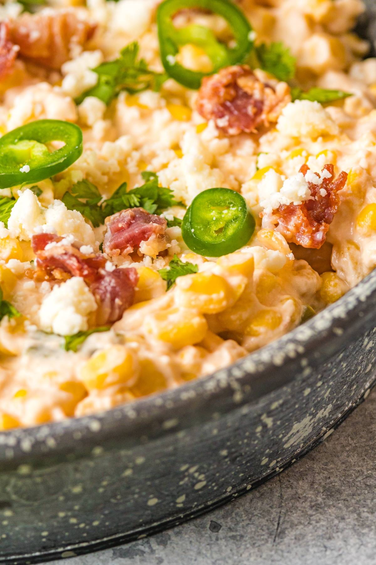 Creamy jalapeño corn dip topped with bacon bits, cilantro, and crumbled cheese in a speckled pan—a cheesy Mexican-style appetizer perfect for sharing.
