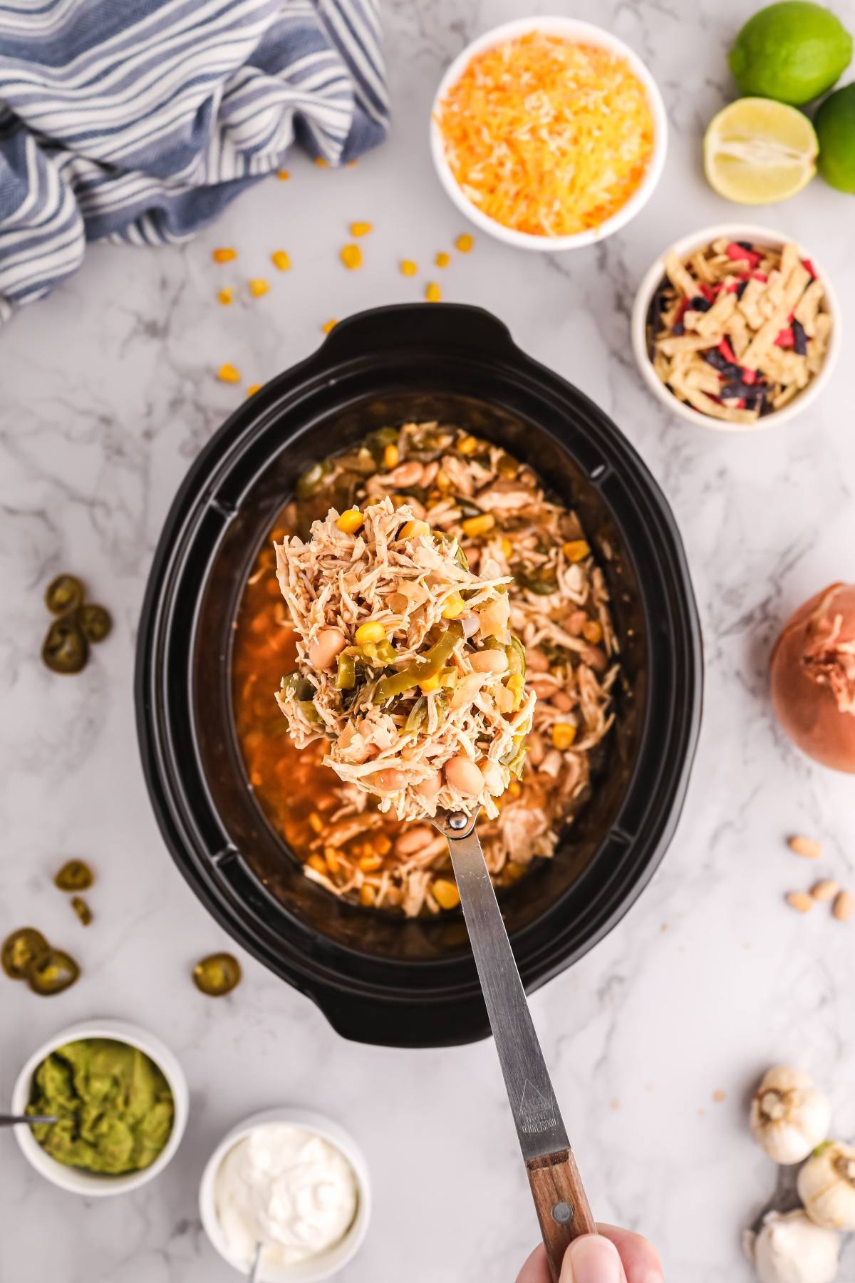 A spoon scoops tender Slow Cooker White Chicken Chili—filled with shredded chicken and veggies—from a crockpot, surrounded by toppings on a marble counter.