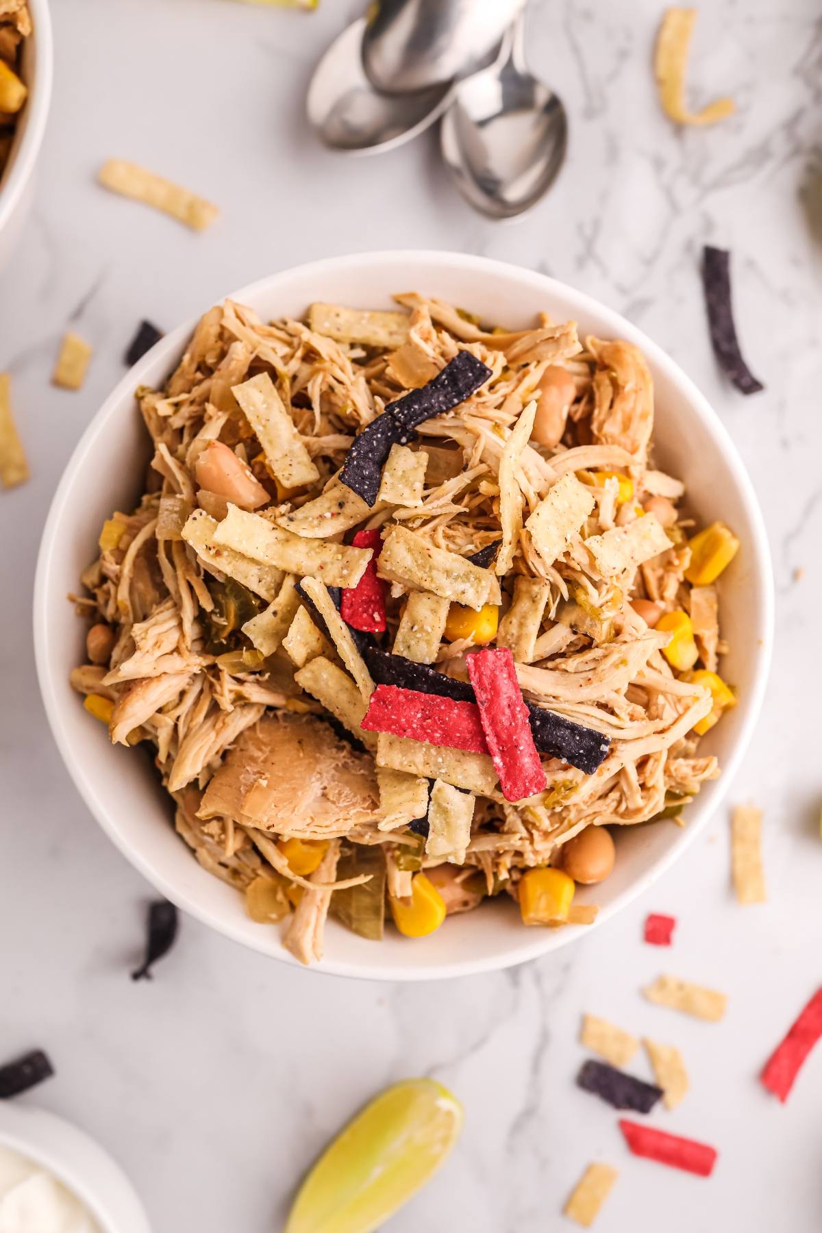 A bowl of shredded chicken, corn, beans, and tortilla strips sits on a marble surface with spoons nearby—perfect for serving up your favorite white bean and chicken chili or trying a new chicken chili recipe.