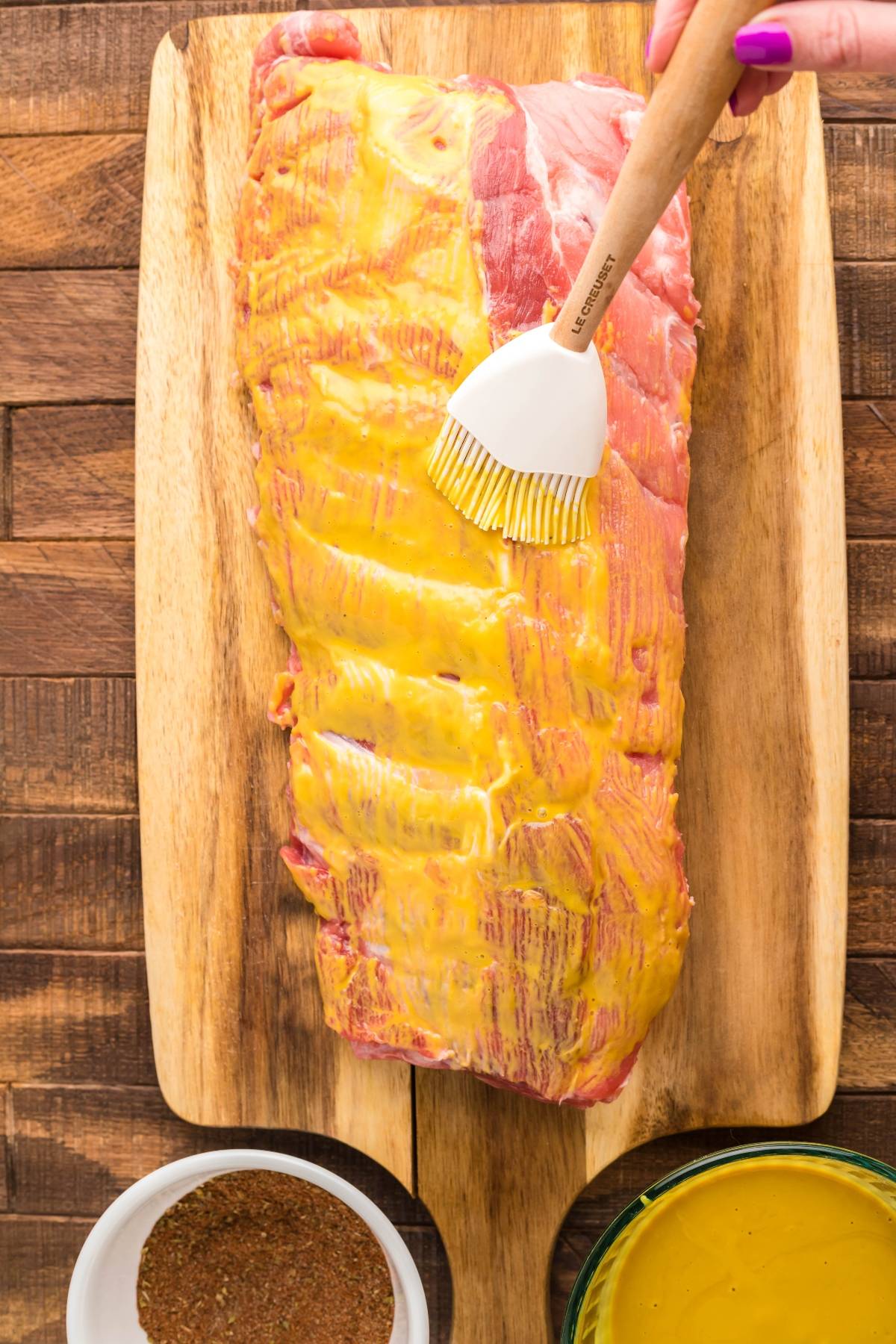 A hand brushes yellow mustard onto a rack of raw smoked baby back ribs on a wooden cutting board, with seasoning nearby.