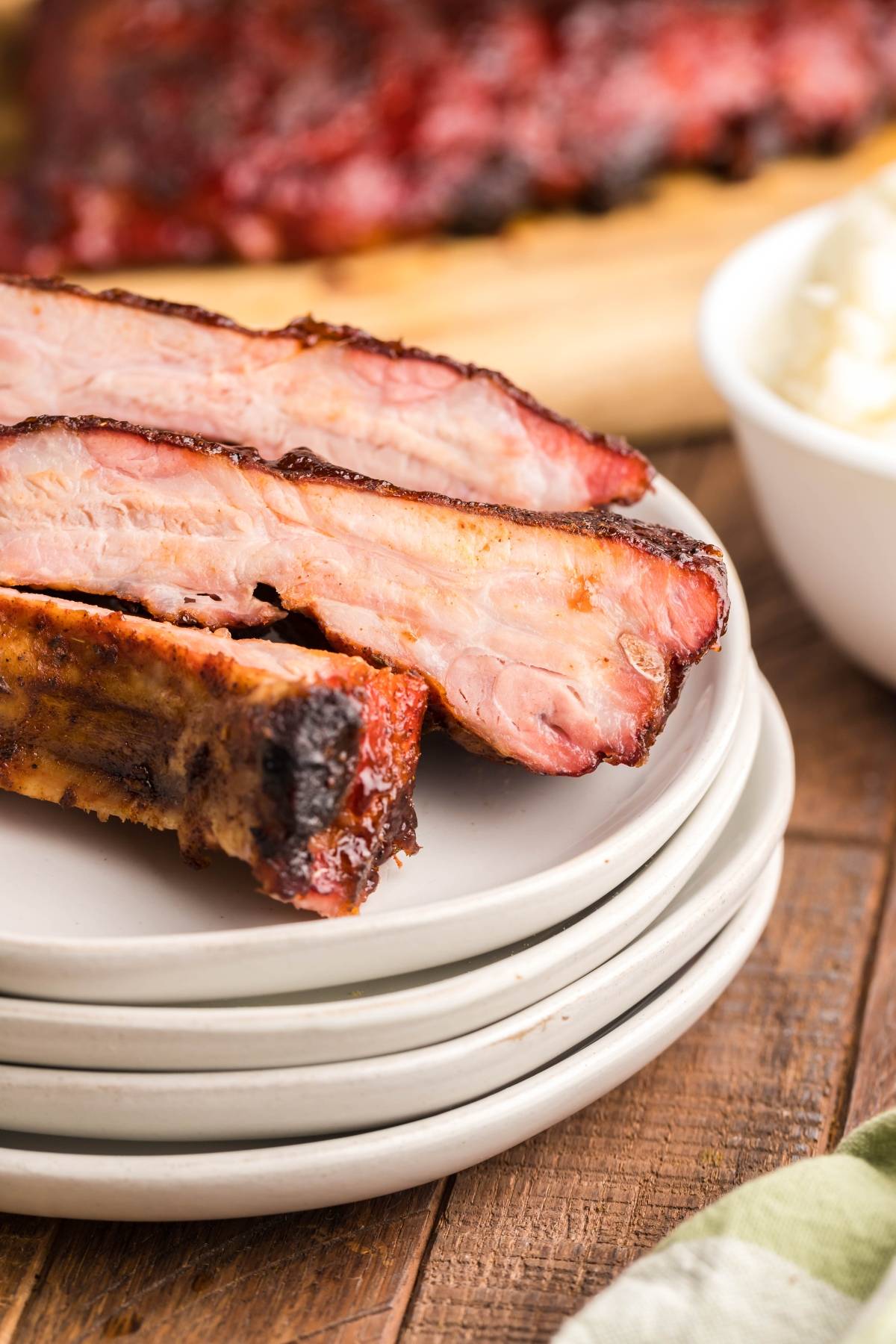 Close-up of three smoked baby back ribs stacked on white plates, with a bowl and more ribs in the background.