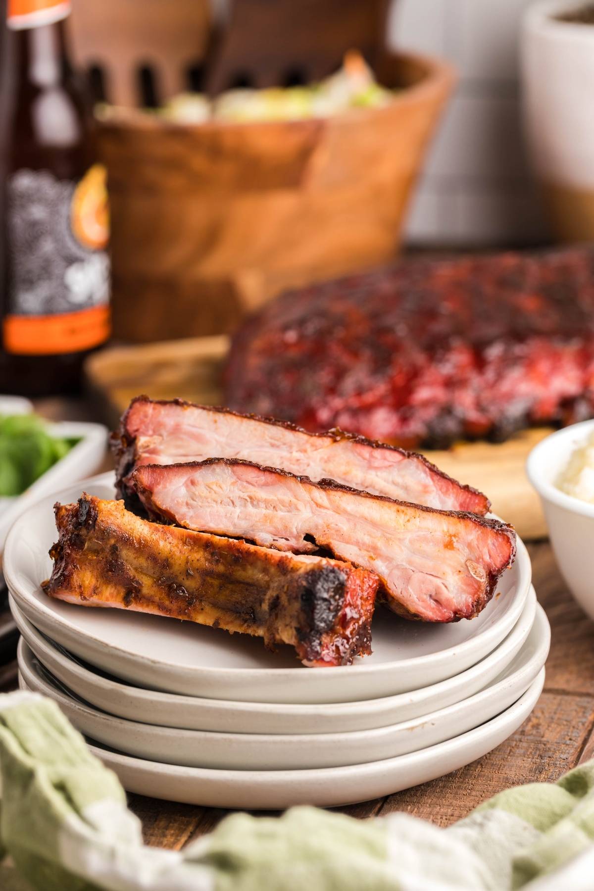 Three smoked baby back ribs stacked on white plates, with a slab of ribs and side dishes in the background—an irresistible Summer BBQ favourite made in a Traeger grill.
