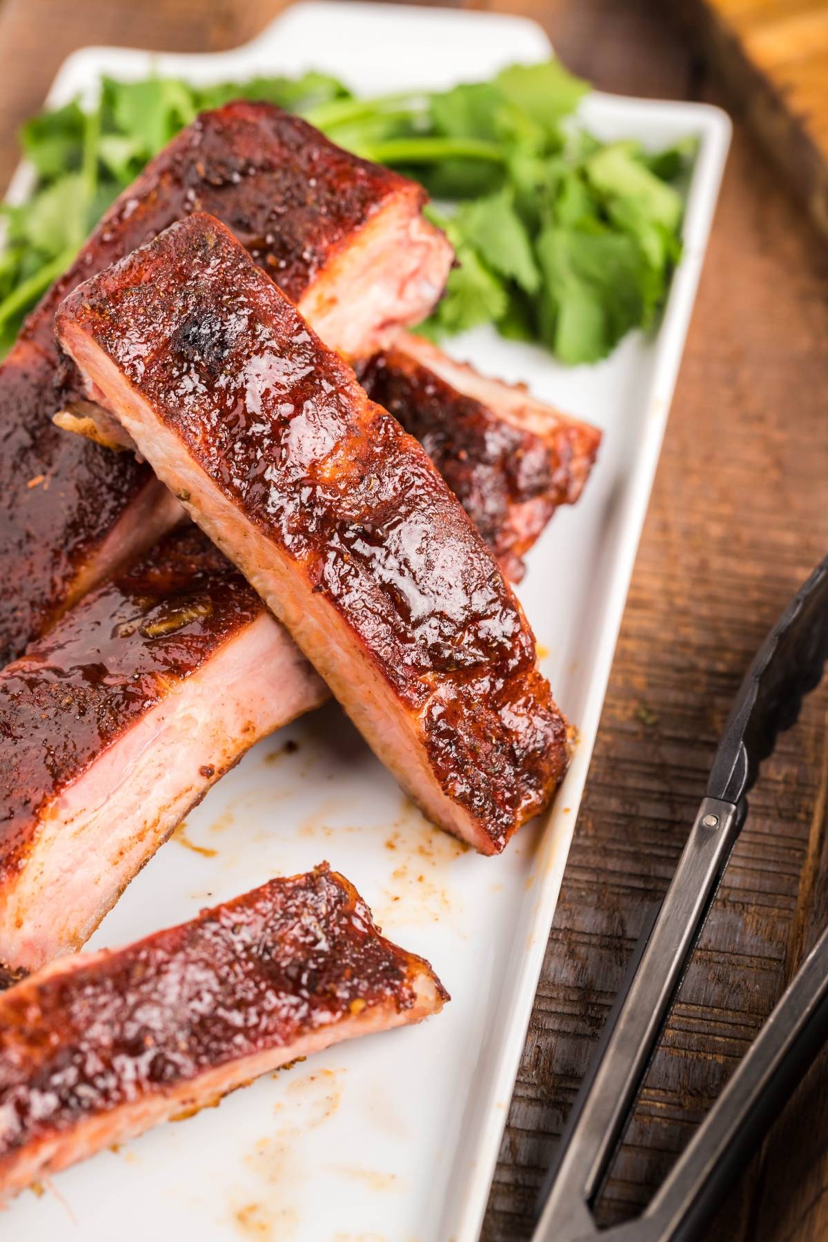 Smoked baby back ribs with a glossy glaze on a white plate, garnished with fresh greens—a true Summer BBQ favourite.