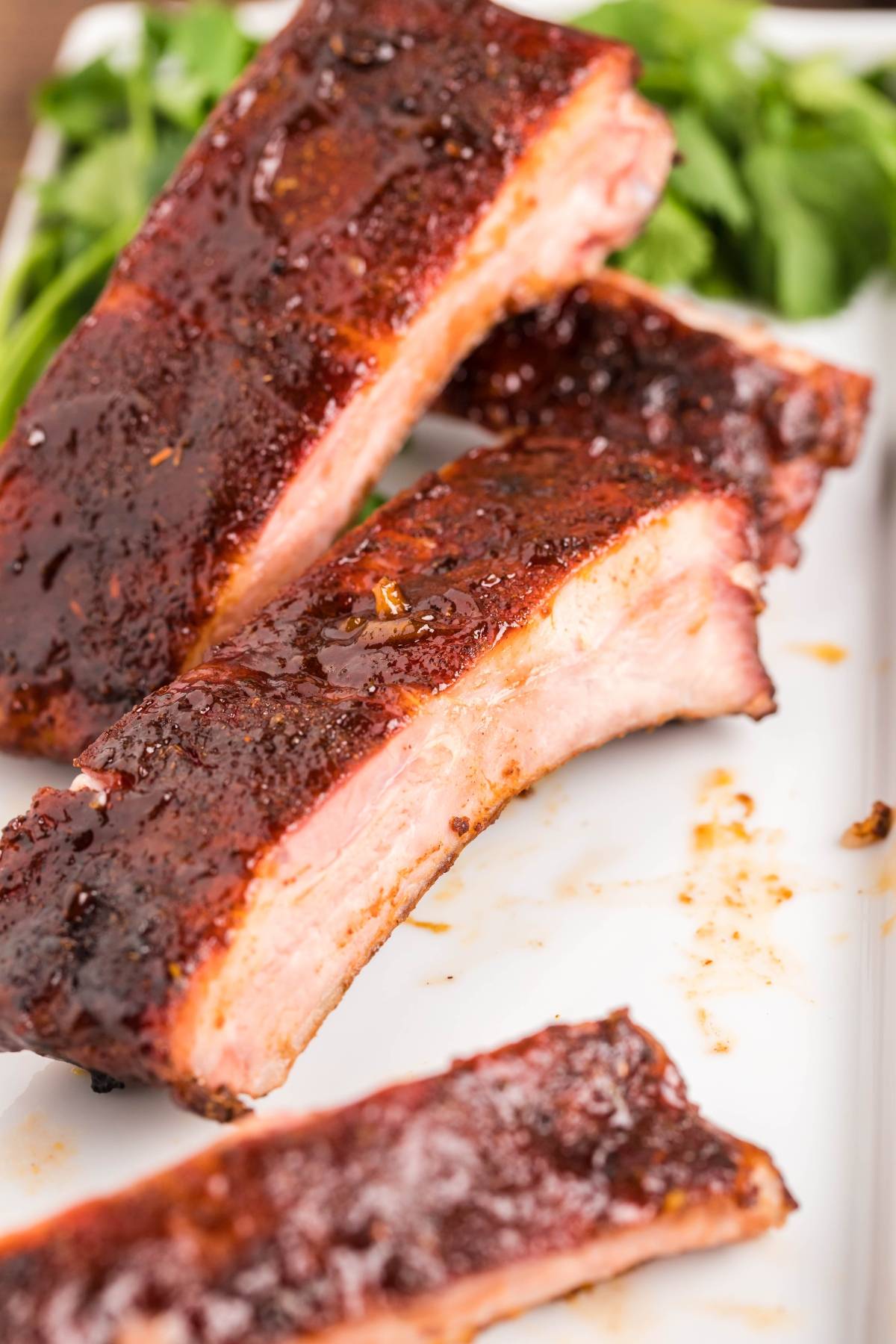 Close-up of juicy, smoked ribs made in a Traeger grill on a white plate with fresh greens in the background.