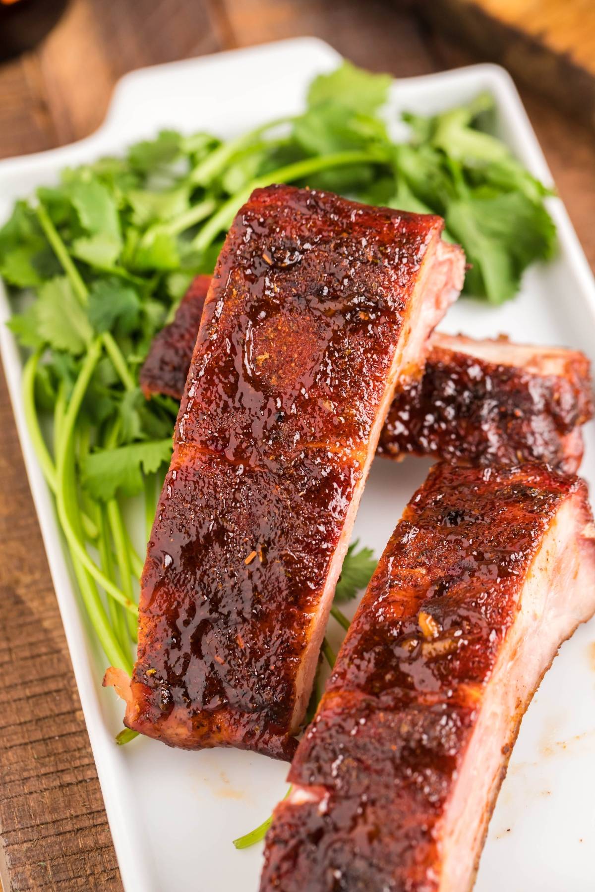 Three glazed smoked baby back ribs on a white plate with fresh cilantro in the background—an easy grilling recipe perfect for any occasion.