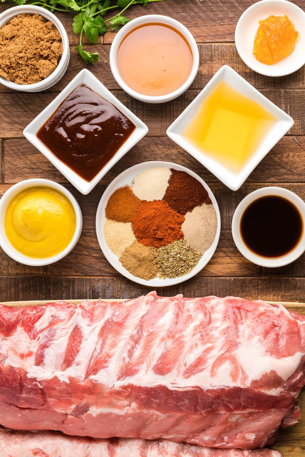 Raw ribs surrounded by various spices, sauces, mustard, brown sugar, and seasonings on a wooden table—perfect for an easy grilling recipe or smoked baby back ribs made in a Traeger grill.