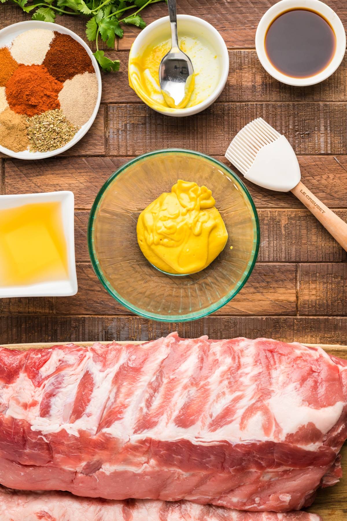 Raw pork ribs with mustard, spices, apple cider vinegar, and sauce in bowls, plus a basting brush on a wooden table—perfect ingredients for a classic BBQ rib recipe or smoked baby back ribs.