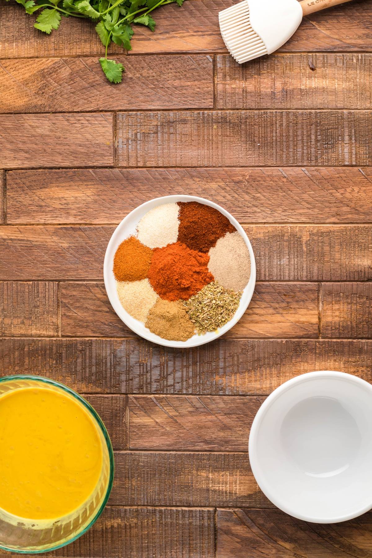 A small plate with assorted spices on a wooden table, surrounded by herbs, a sauce, a brush, and an empty bowl—perfect for prepping smoked baby back ribs or your favorite easy grilling recipe.