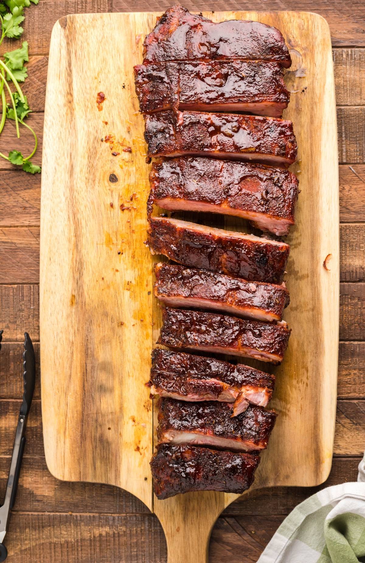 Sliced barbecued ribs on a wooden cutting board, with tongs and herbs nearby—an easy grilling recipe perfect for anyone craving smoked ribs made in a Traeger grill.