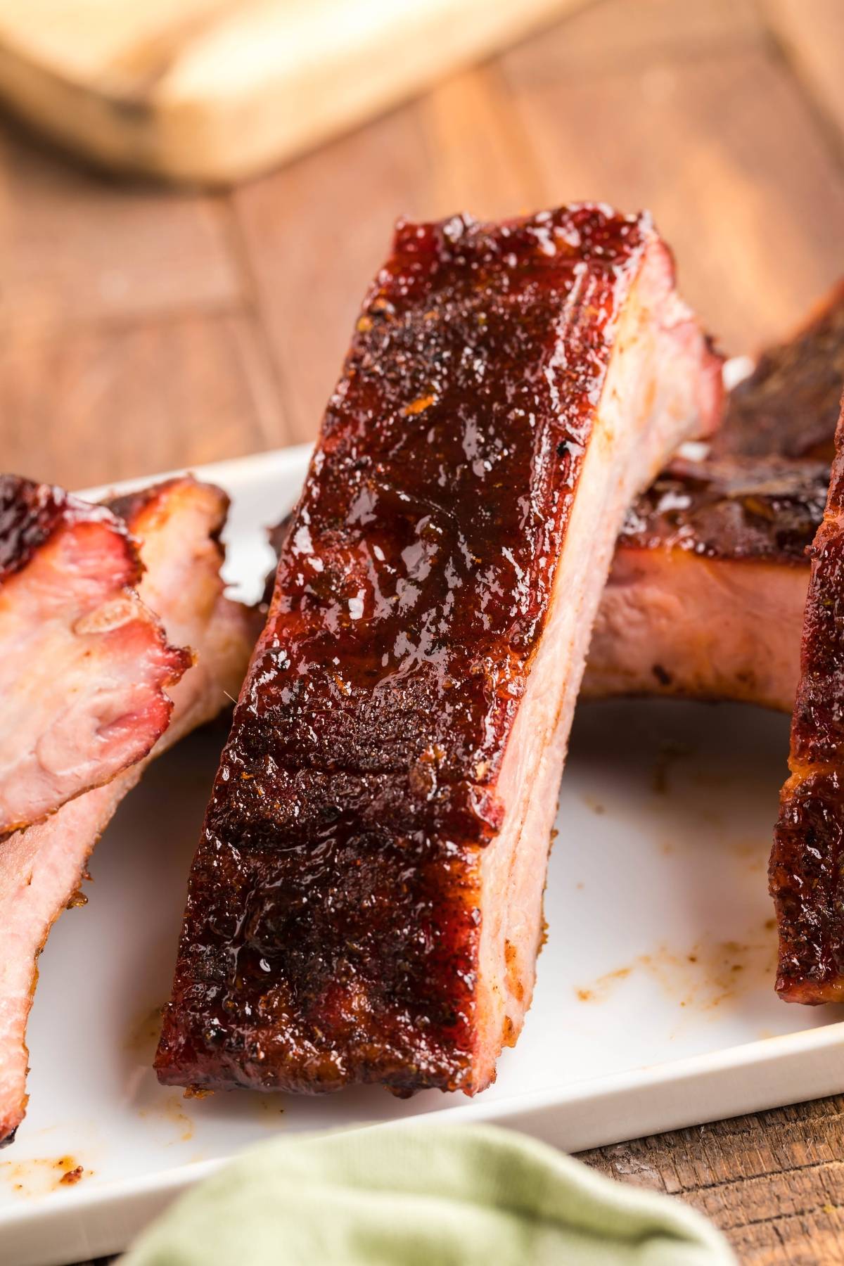 Close-up of glazed, juicy smoked baby back ribs stacked on a white plate, with a wooden table in the background—a mouthwatering showcase of this BBQ rib recipe.