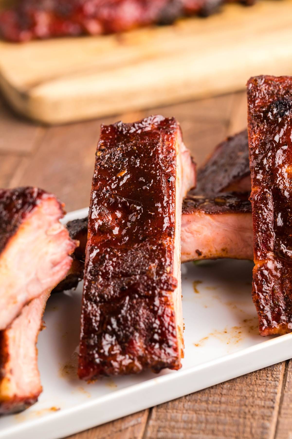 Close-up of saucy smoked baby back ribs stacked on a white plate, highlighting a delicious BBQ rib recipe, with a wooden board in the background.