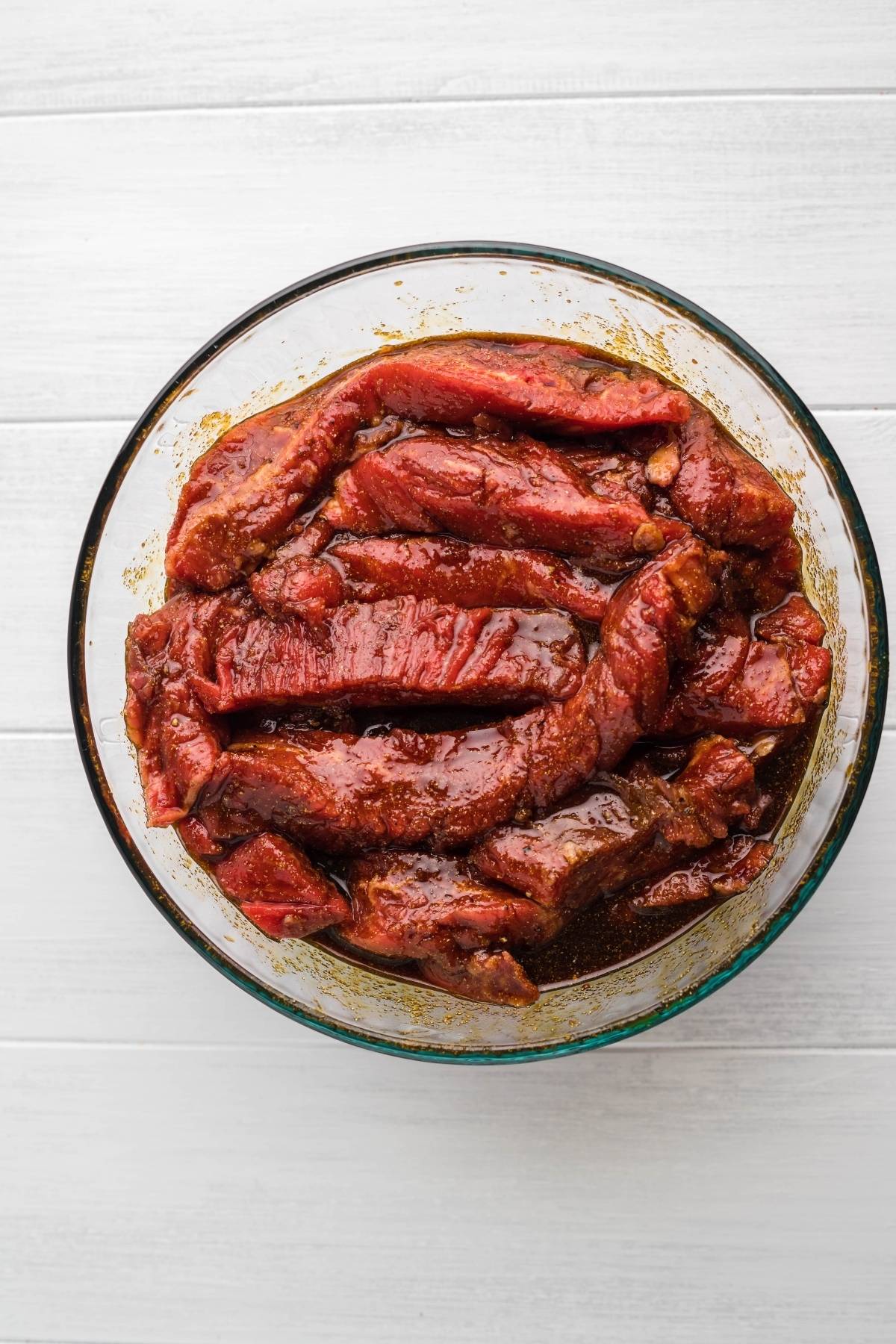 Raw beef strips marinating in a glass bowl with spices, ready to be transformed into healthy homemade beef jerky on a white wooden surface.