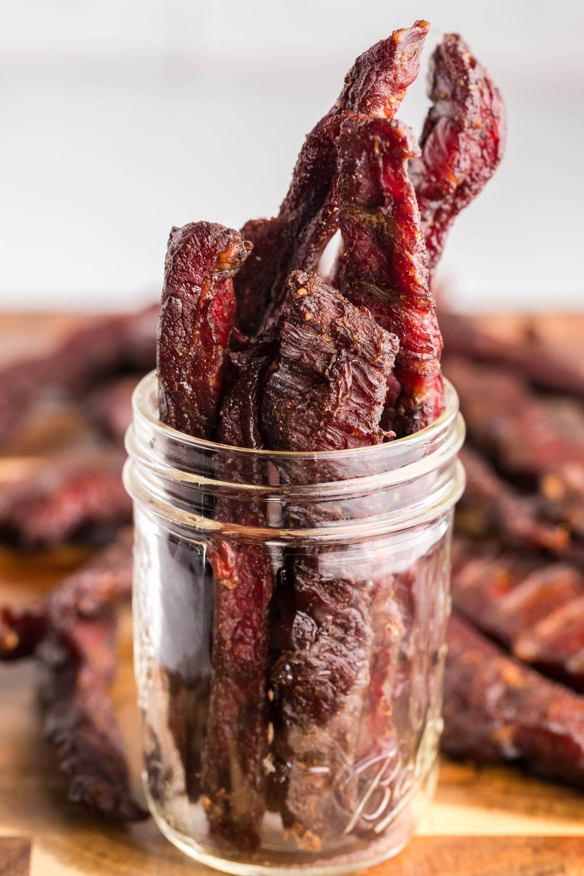 Several pieces of gluten free paleo jerky stand upright in a clear glass jar, with more jerky in the background—perfect inspiration if you’re searching for a recipe for a smoker like a Traeger grill.