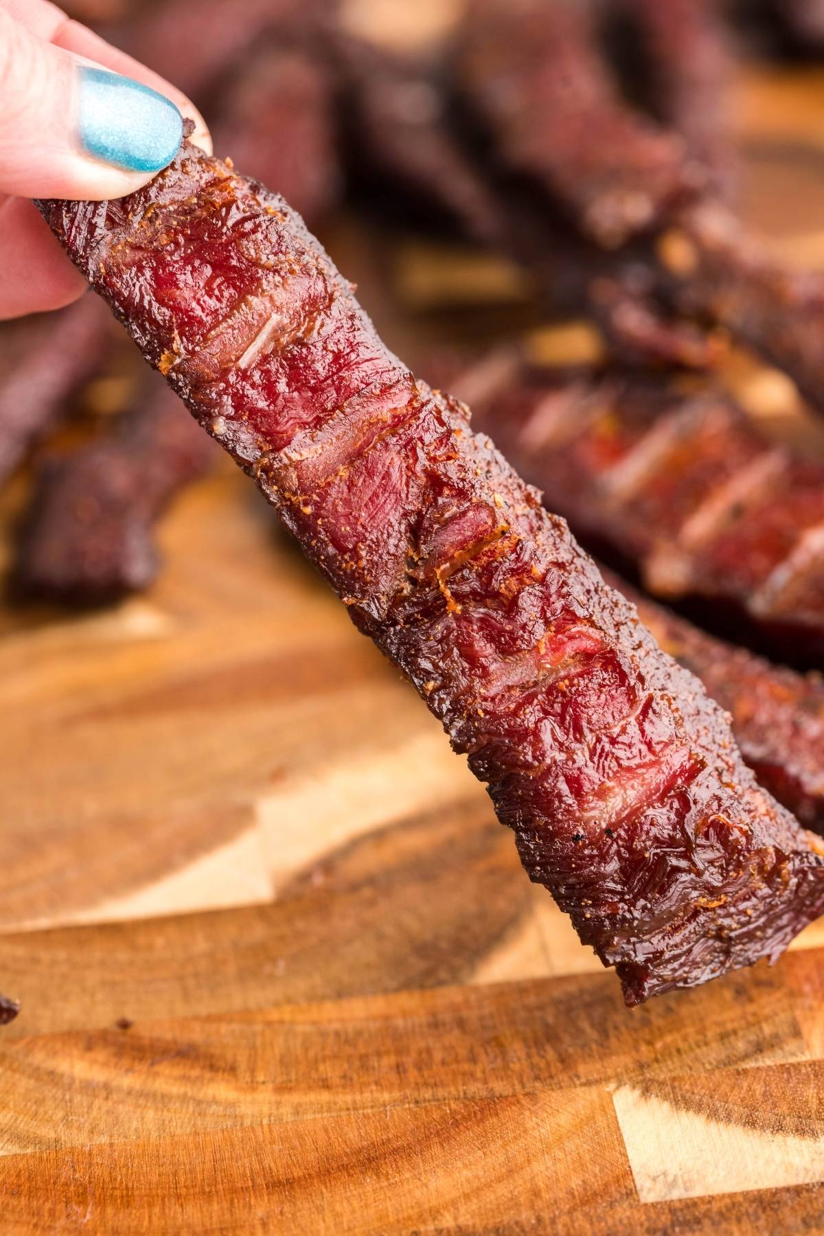 A hand holding a piece of smoked beef jerky over a wooden cutting board, with more jerky in the background—perfect for those looking for how to make smoked beef jerky or try a recipe for a smoker like a Traeger grill.