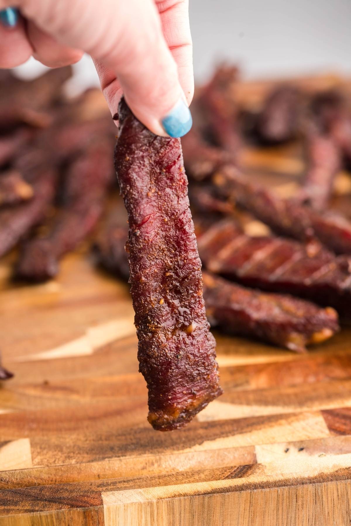 A hand holds a piece of healthy homemade beef jerky above a wooden board topped with more smoked beef jerky pieces.