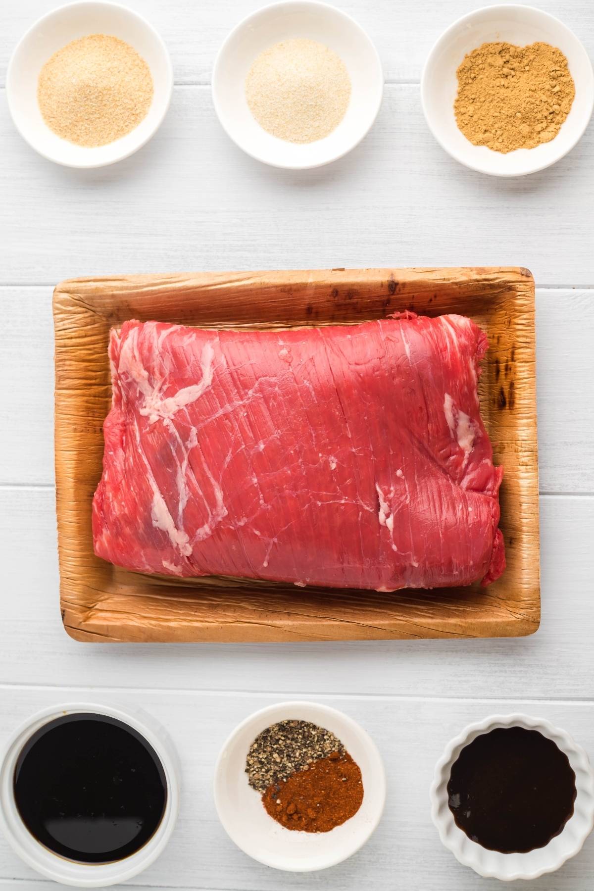 Raw flank steak on a tray, surrounded by bowls of spices and sauces—perfect for healthy homemade beef jerky or trying a recipe for a smoker like a Traeger grill.