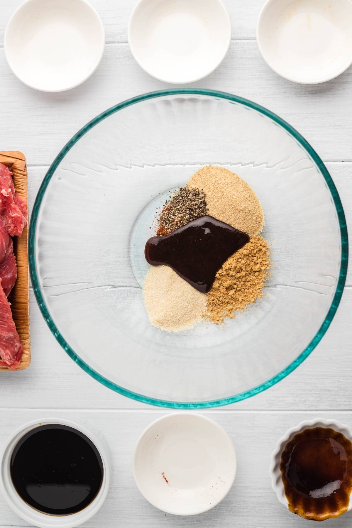 A glass bowl with spices and sauce, surrounded by small bowls and raw meat slices on a white surface—perfect for making healthy homemade beef jerky.