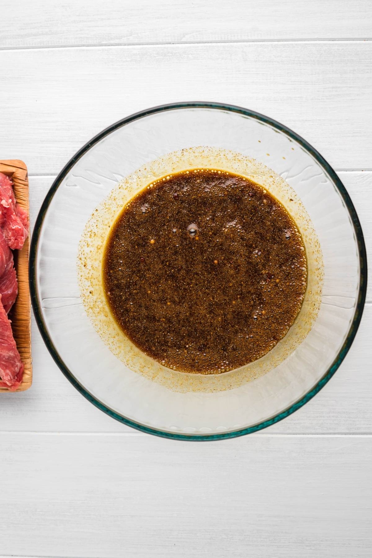 Large glass bowl of dark brown marinade next to a wooden tray with pieces of raw beef on a white surface—perfect for making smoked beef jerky or gluten free paleo jerky using your favorite recipe for a smoker like a Traeger grill.