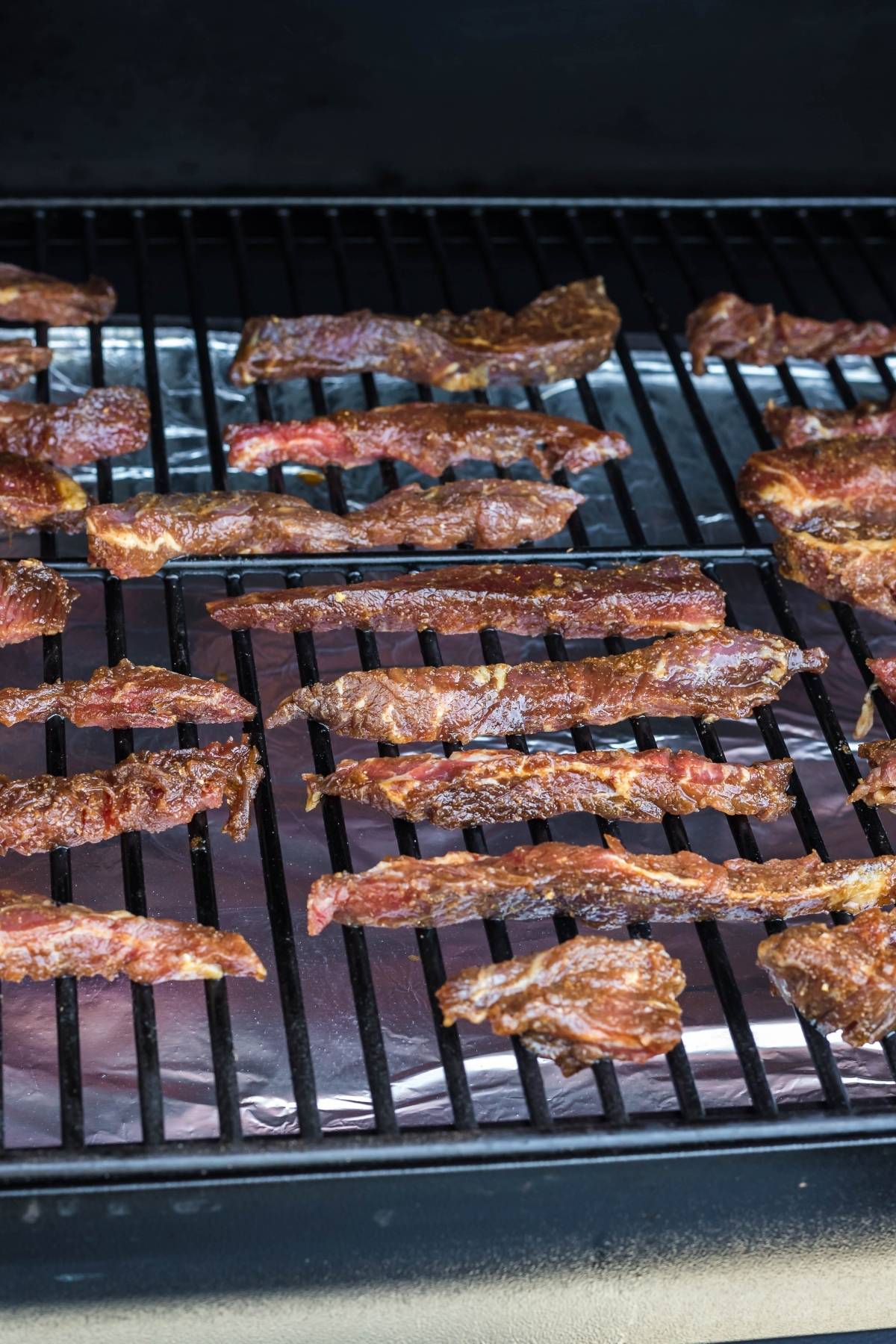 Thin strips of healthy homemade beef jerky being smoked on a metal grill rack covered with foil underneath—perfect for a gluten free paleo jerky recipe for a smoker like a Traeger grill.