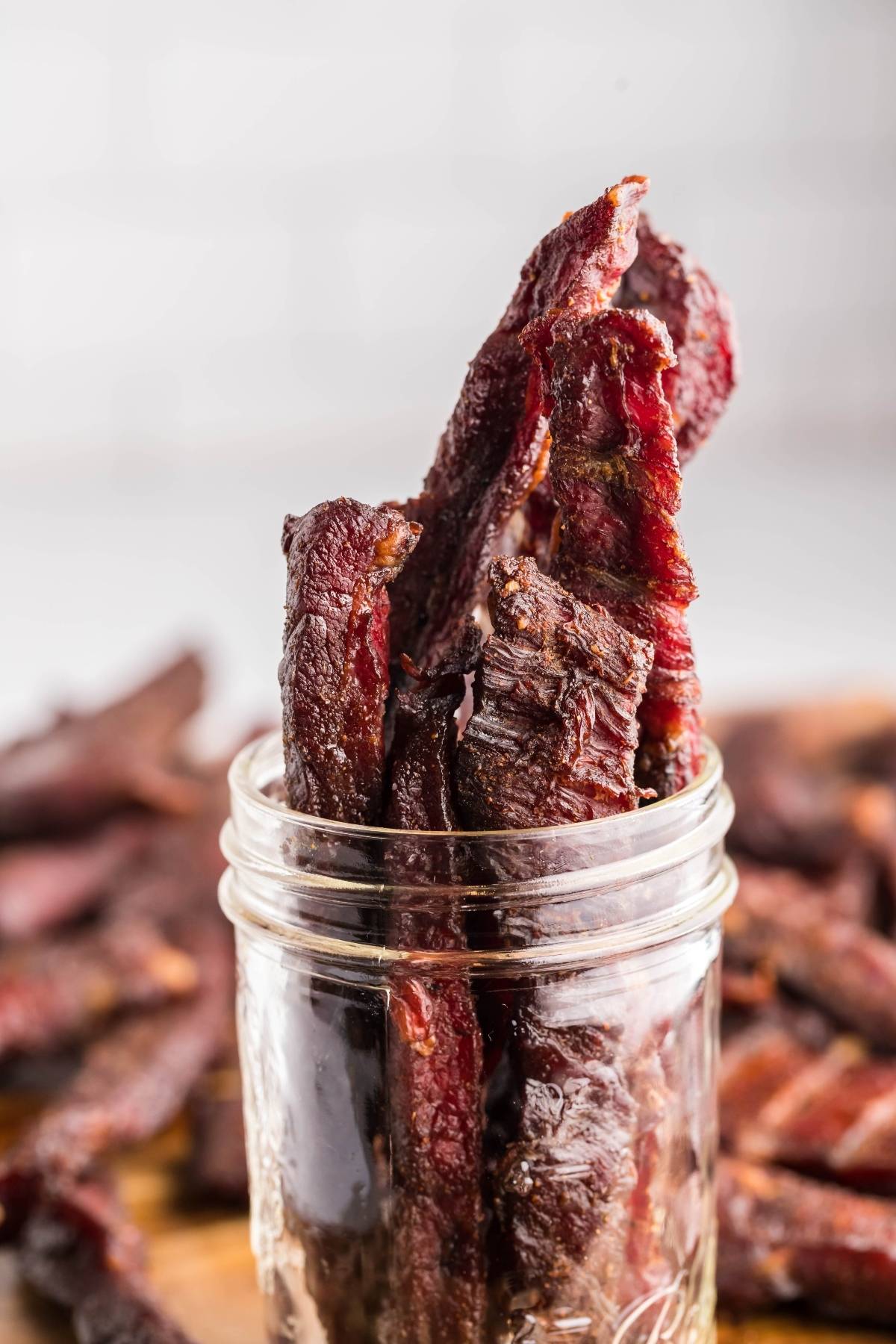 Several pieces of smoked beef jerky stand upright in a clear glass jar, with more gluten free paleo jerky visible in the background.
