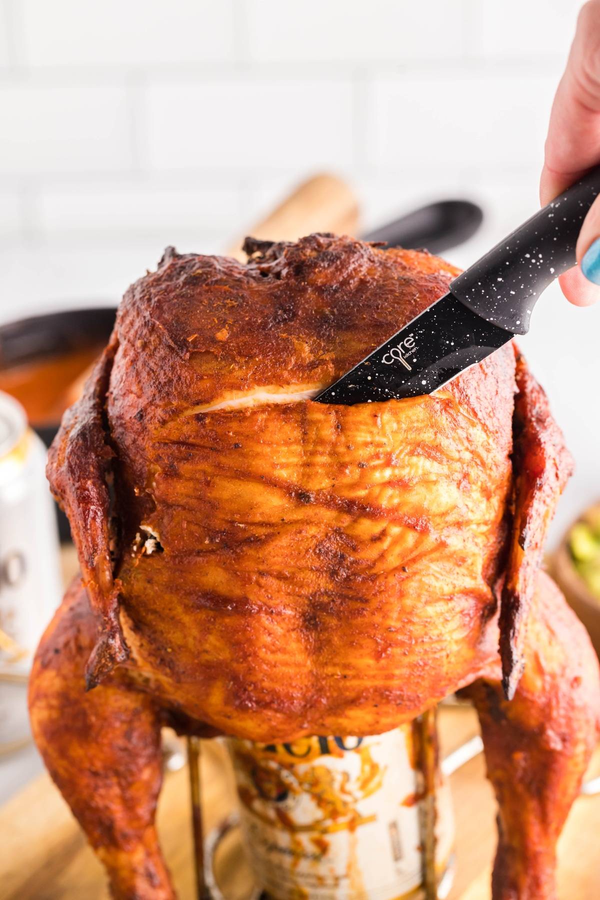 A knife slices into a roasted Traeger beer can chicken standing upright on an open can.