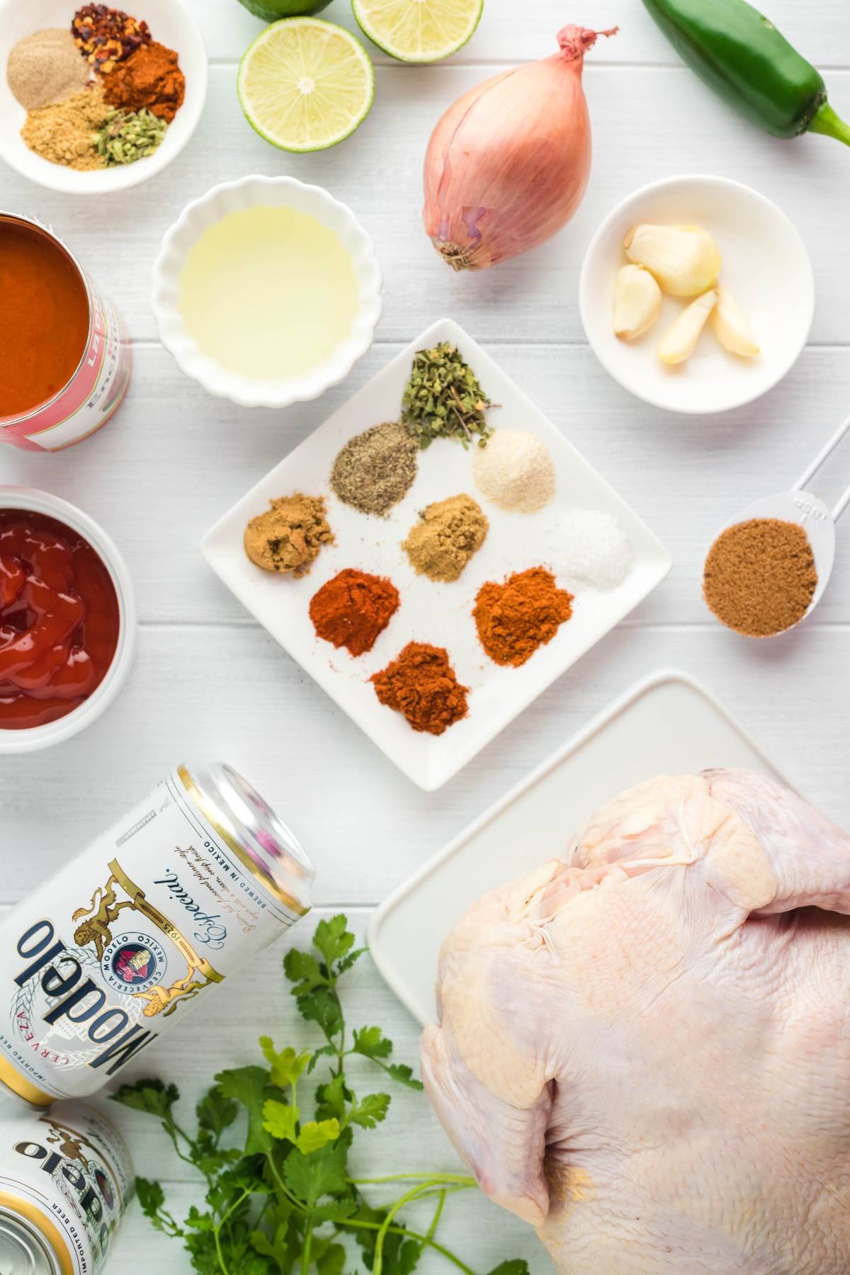 Raw whole chicken, assorted spices, fresh herbs, canned ingredients, lime, shallot, garlic, and beer on a table—everything you need to make beer can chicken for the perfect grilled chicken recipe.