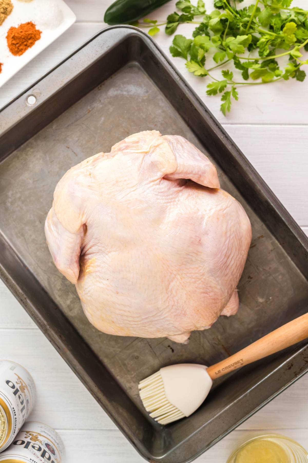 Raw whole chicken in a baking tray with a basting brush, herbs, spices, and cans nearby on a white table—perfect setup to make beer can chicken or try smoked beer can chicken recipes for extra flavor.