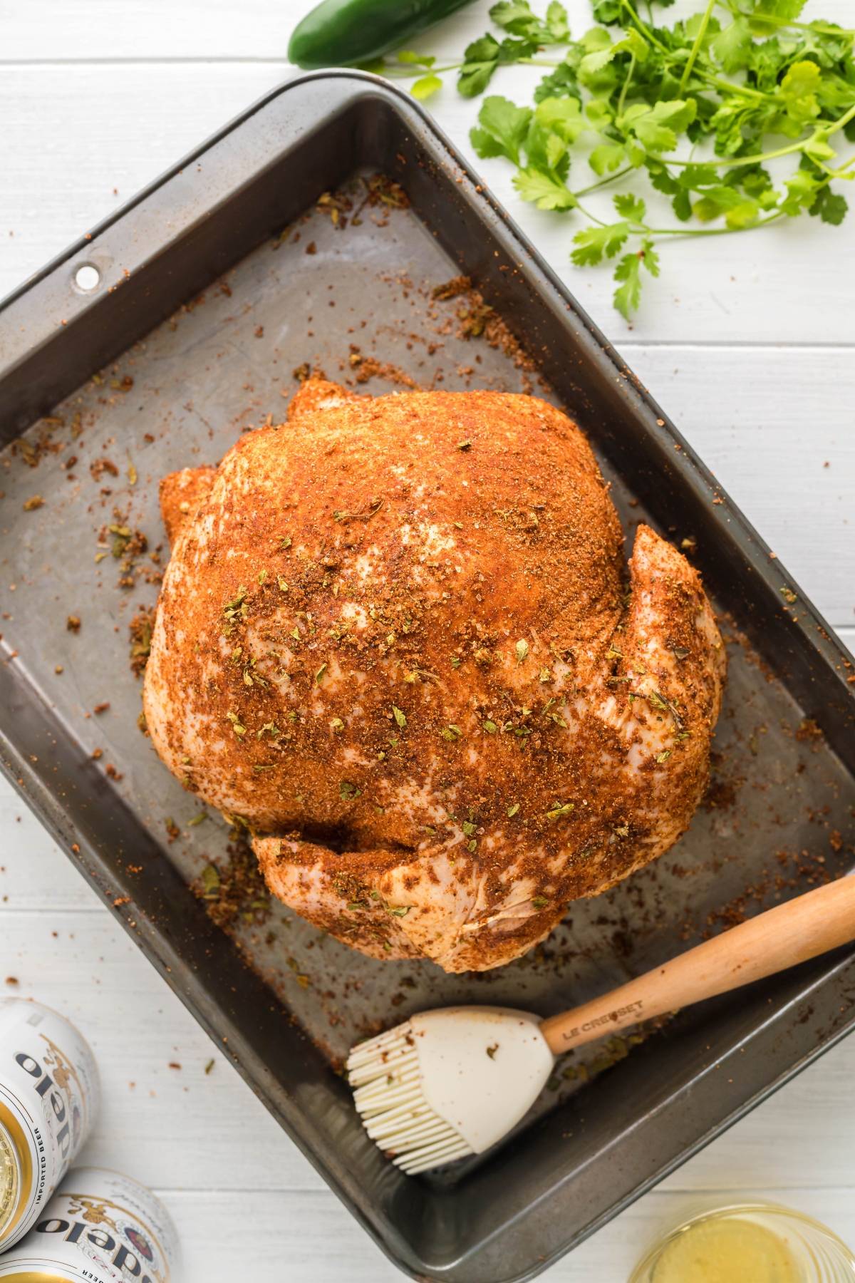 Whole raw chicken coated with spices in a roasting pan, surrounded by herbs, a brush, and canned drinks—perfect to make beer can chicken for juicy, flavorful results.