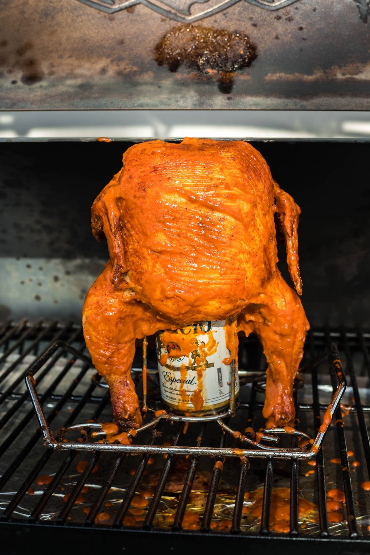 Whole seasoned chicken roasting upright on a grill, sitting on a beer can with a metal stand—try this Traeger beer can chicken for an irresistible grilled chicken recipe.