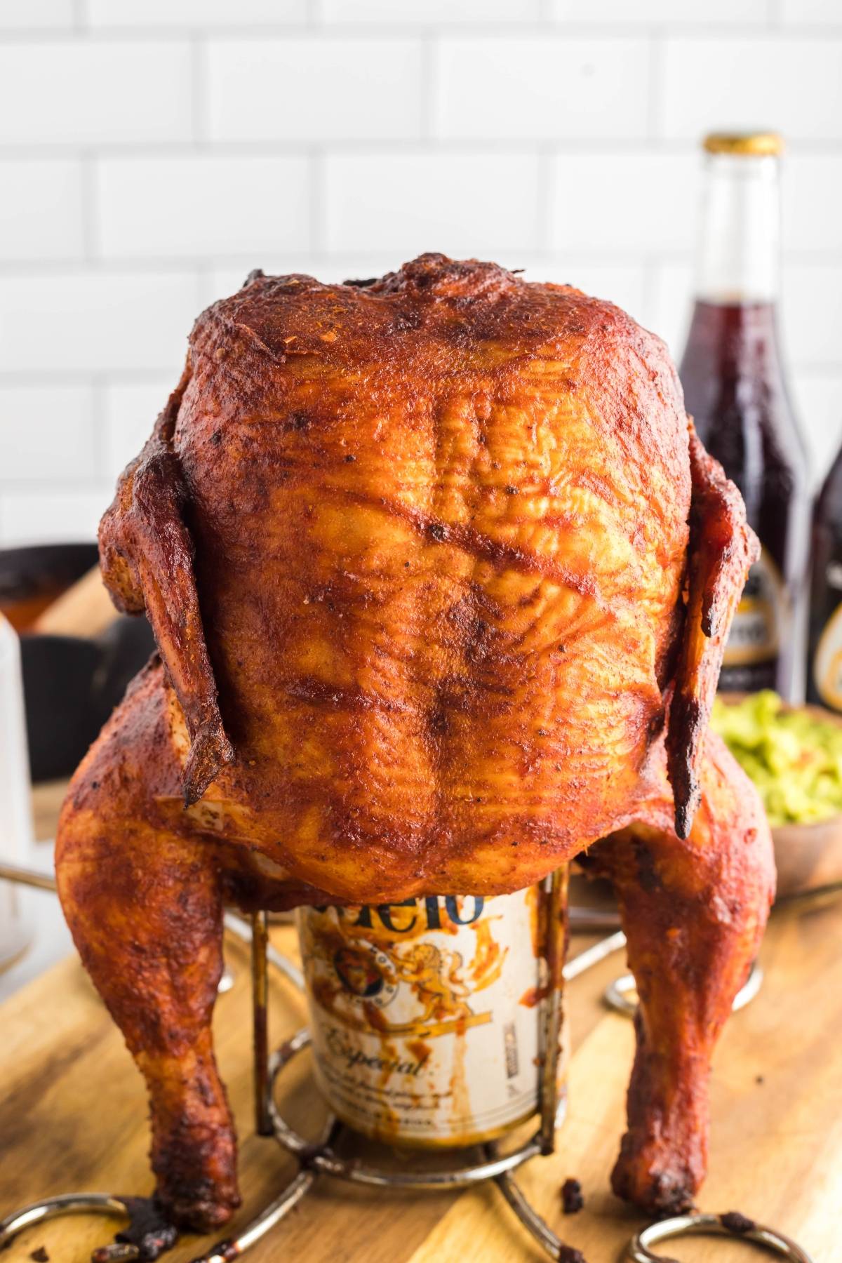 A whole roasted chicken upright on a beer can—perfect for your Traeger beer can chicken or served with tangy Mexican BBQ sauce—set on a wooden surface with drinks in the background.