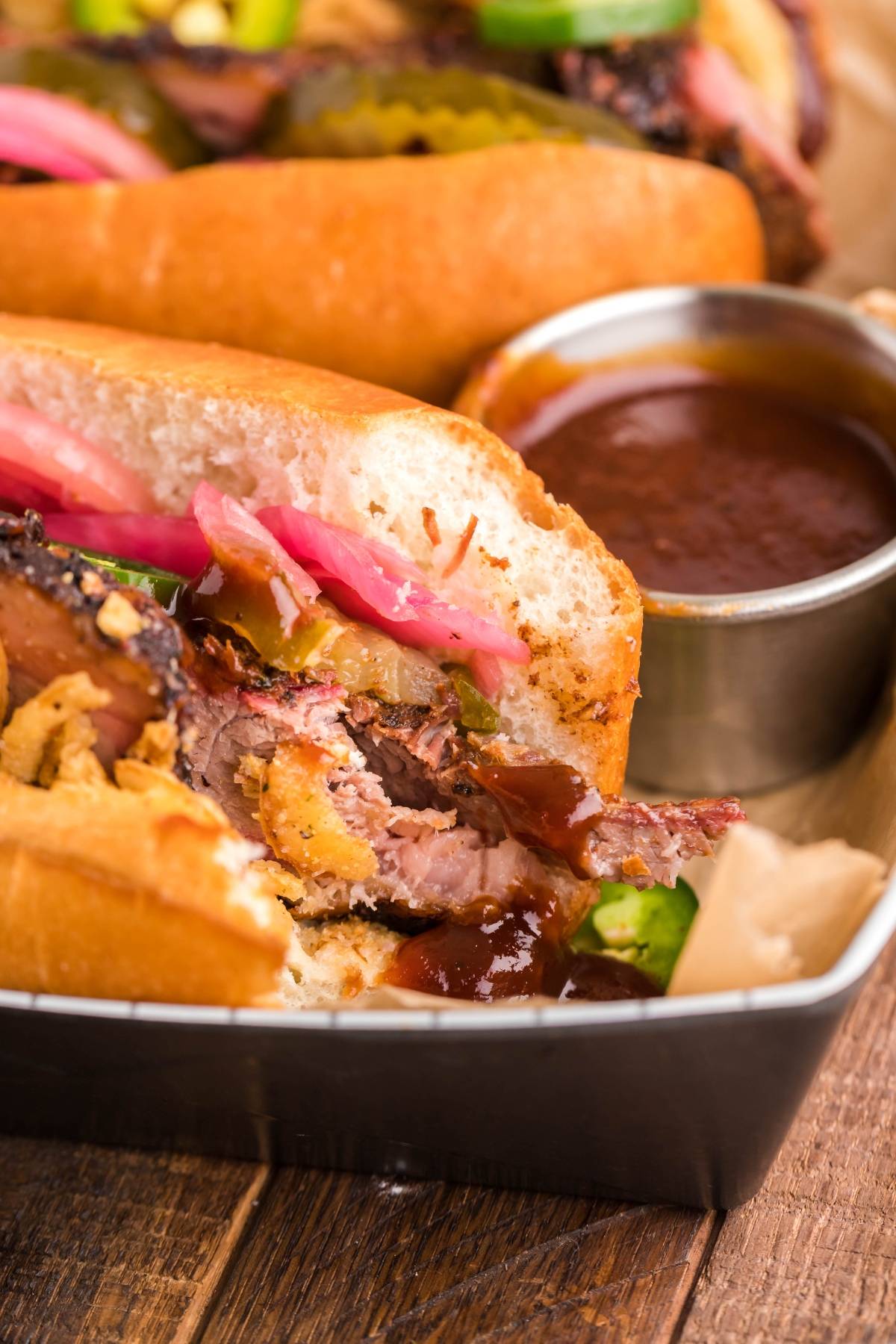 Smoked Brisket Sandwich with pickled onions and classic brisket sandwich toppings, served in a tray with a cup of barbecue sauce on the side.