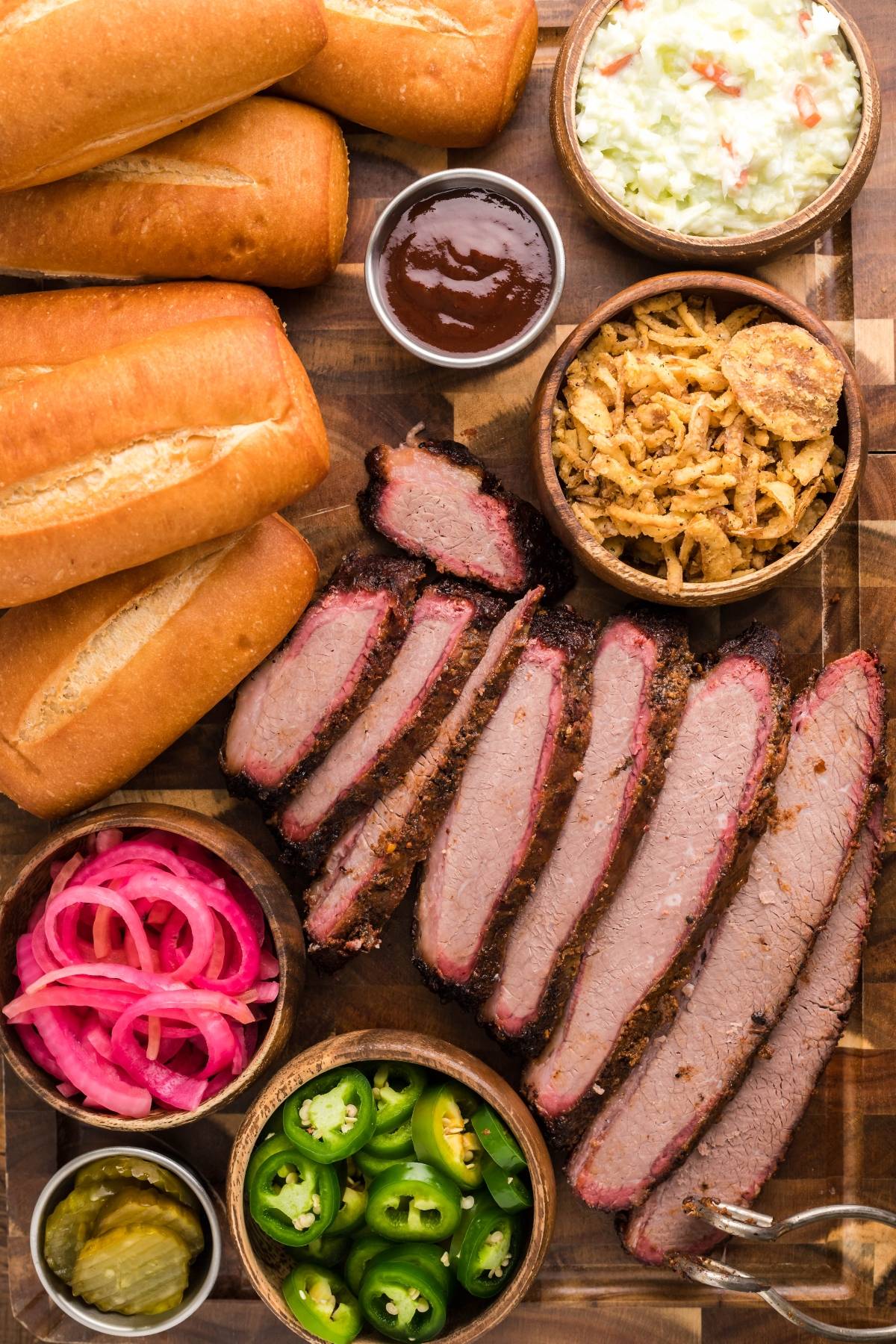 Sliced brisket, hoagie rolls, coleslaw, Homemade Pickled Red Onions, jalapeños, and barbecue sauce on a board—everything you need for the best BBQ brisket sandwich or a tasty sandwich using leftover brisket.