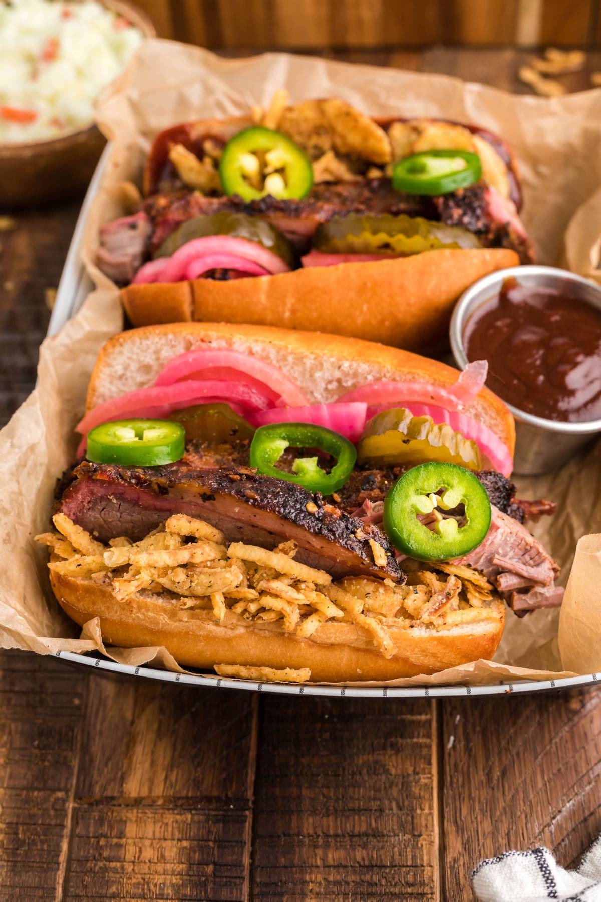 Two BBQ brisket sandwiches with homemade pickled red onions, jalapeños, and crispy onions on a tray with BBQ sauce—a top contender for the best BBQ brisket sandwich and loaded with delicious brisket sandwich toppings.