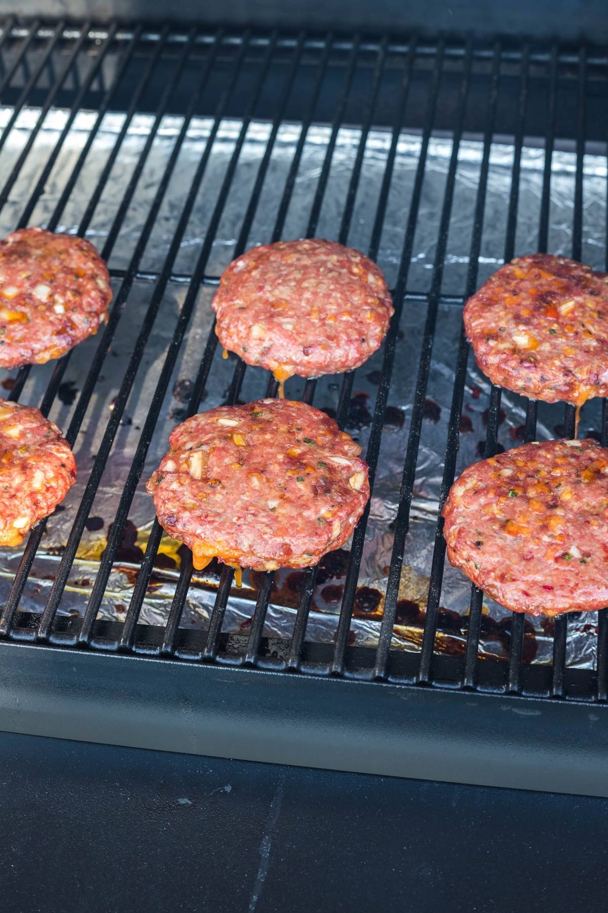 Six raw Smoked Cheddar Bacon Stuffed Burgers cooking on a grill with foil underneath to catch drips.