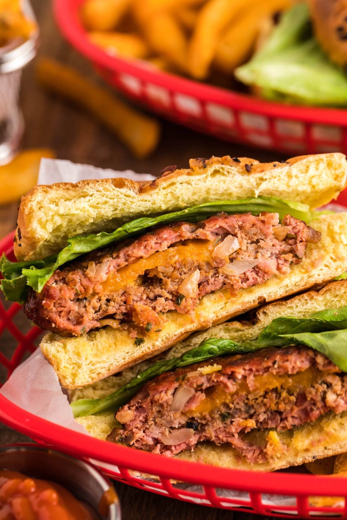 A close-up of a Smoked Cheddar Bacon Stuffed Burger cut in half, topped with lettuce, in a red basket with fries in the background.
