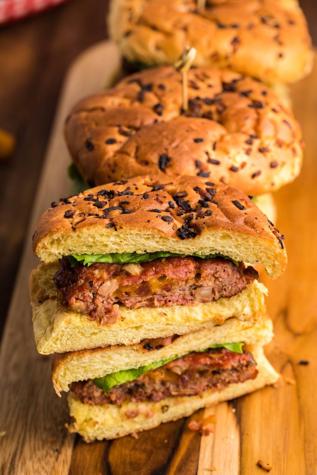 Stacked bacon and cheese stuffed burgers with lettuce on onion buns, sliced in half and served on a wooden board.