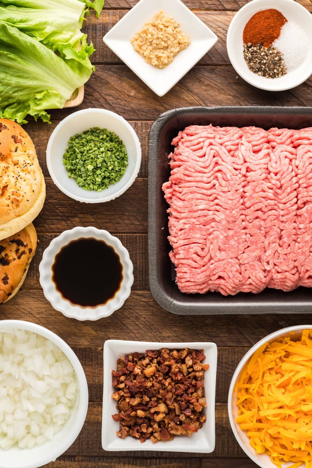 Top-down view of Smoked Cheddar Bacon Stuffed Burgers ingredients: ground beef, lettuce, cheese, onions, bacon, buns, and seasonings on a table.