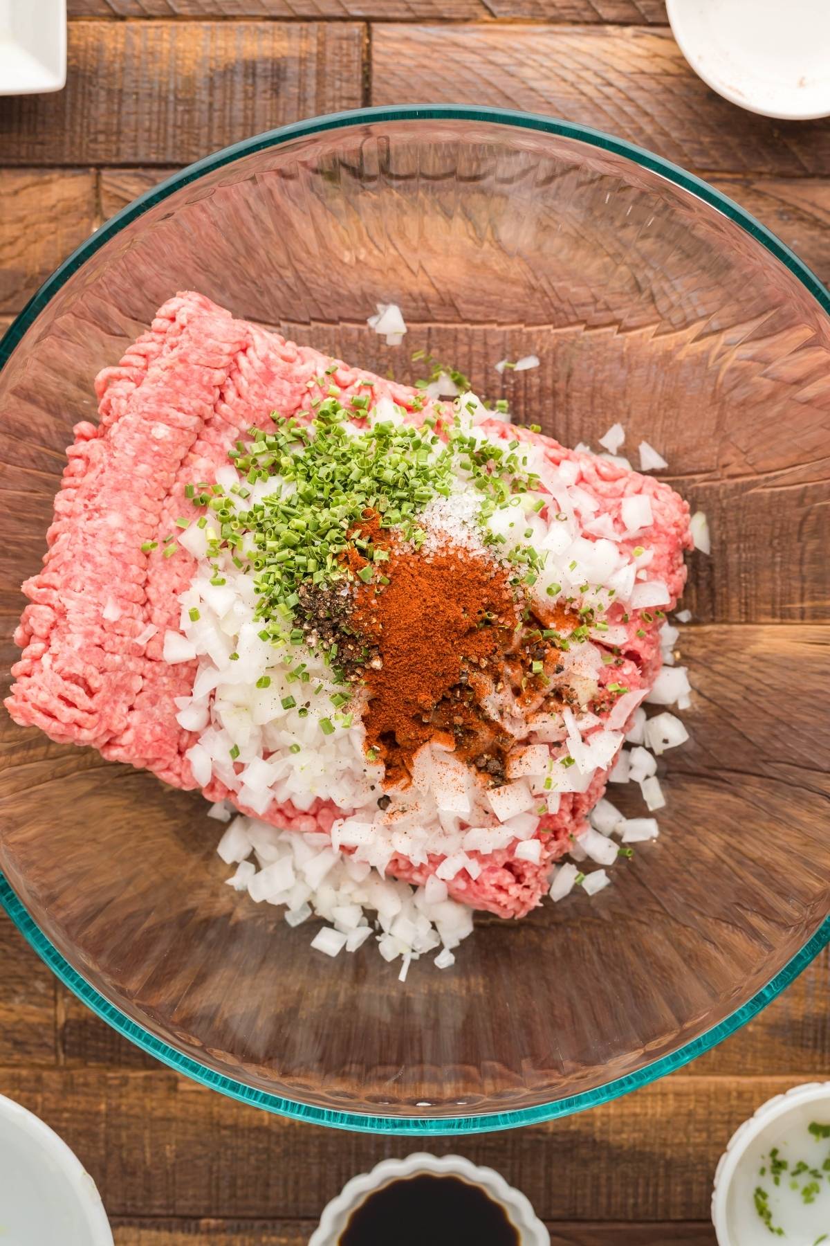 Ground beef, chopped onions, herbs, and spices in a glass bowl on a wooden table, ready to be mixed for delicious smoked or grilled burgers.
