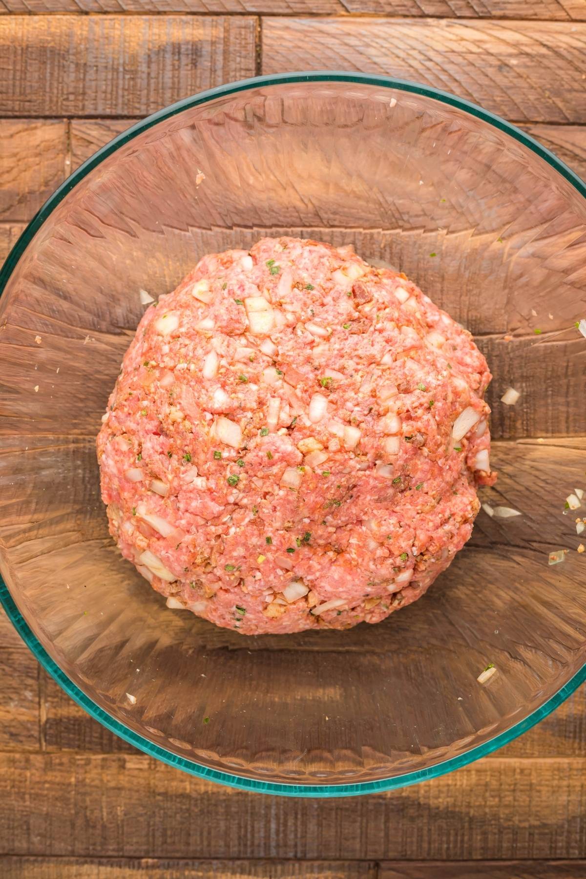 Ground meat mixed with diced onions and herbs in a clear glass bowl on a wooden surface, perfect for creating Traeger grilled stuffed burgers.
