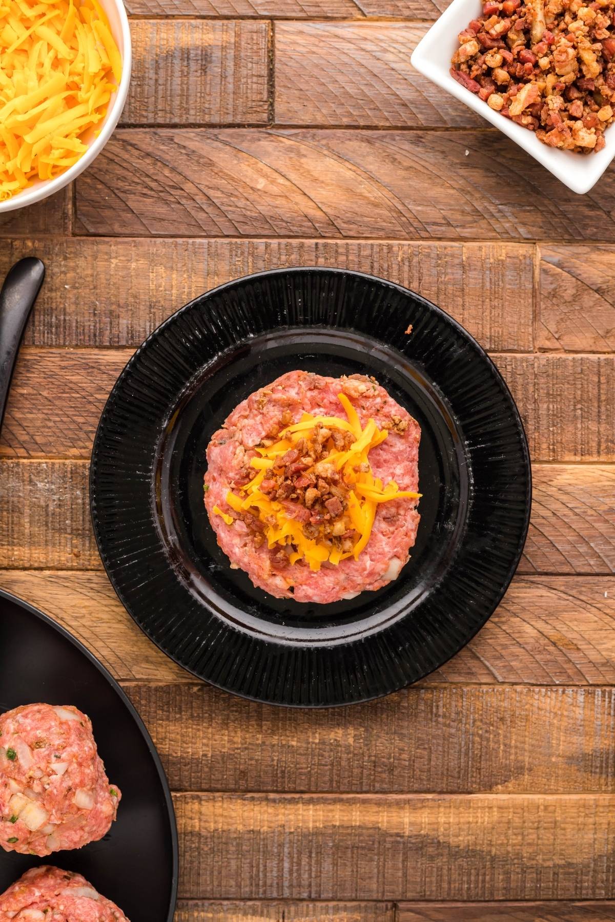 Raw Traeger grilled stuffed burger patty topped with shredded cheese and bacon bits on a black plate, surrounded by ingredients.