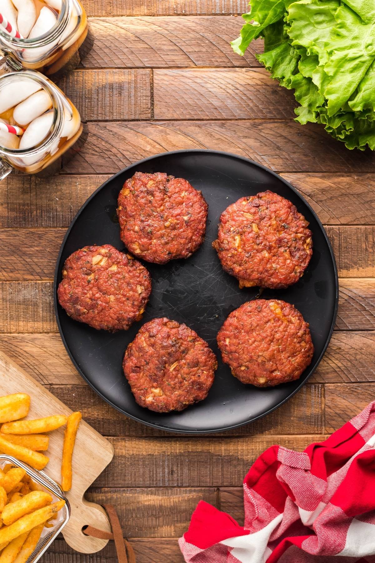 Five uncooked smoked cheddar bacon stuffed burgers on a black plate, with fries, lettuce, drinks, and a red towel nearby.