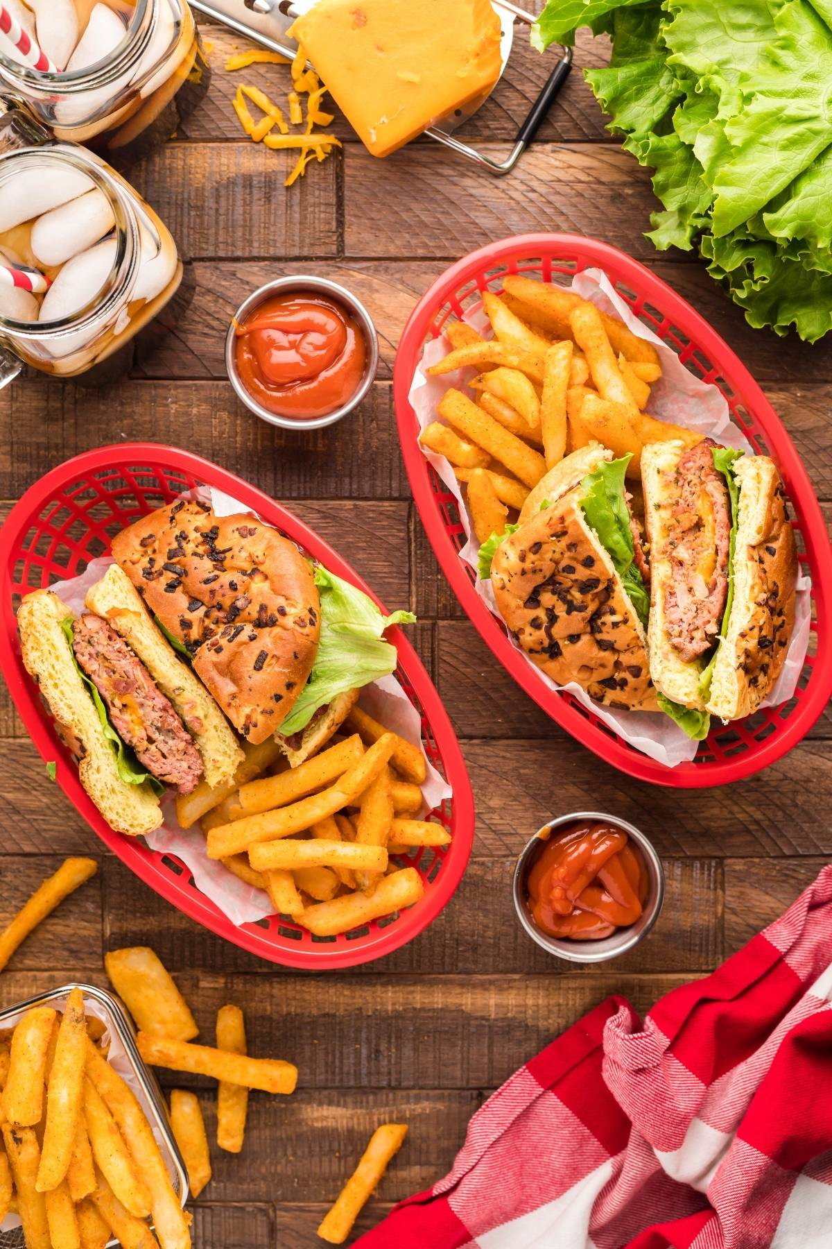 Two smoked Cheddar bacon stuffed burgers with lettuce and fries in red baskets on a wooden table, with ketchup, cheese, and drinks nearby.