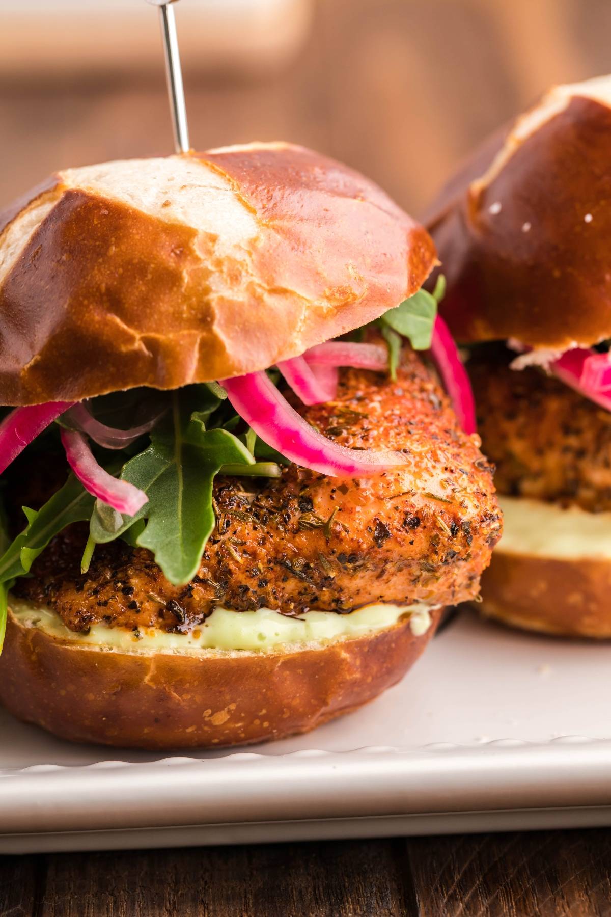 A close-up of a seasoned chicken sandwich with arugula, pickled onions, and sauce on a pretzel bun, made in a pellet grill for extra smoky flavor.