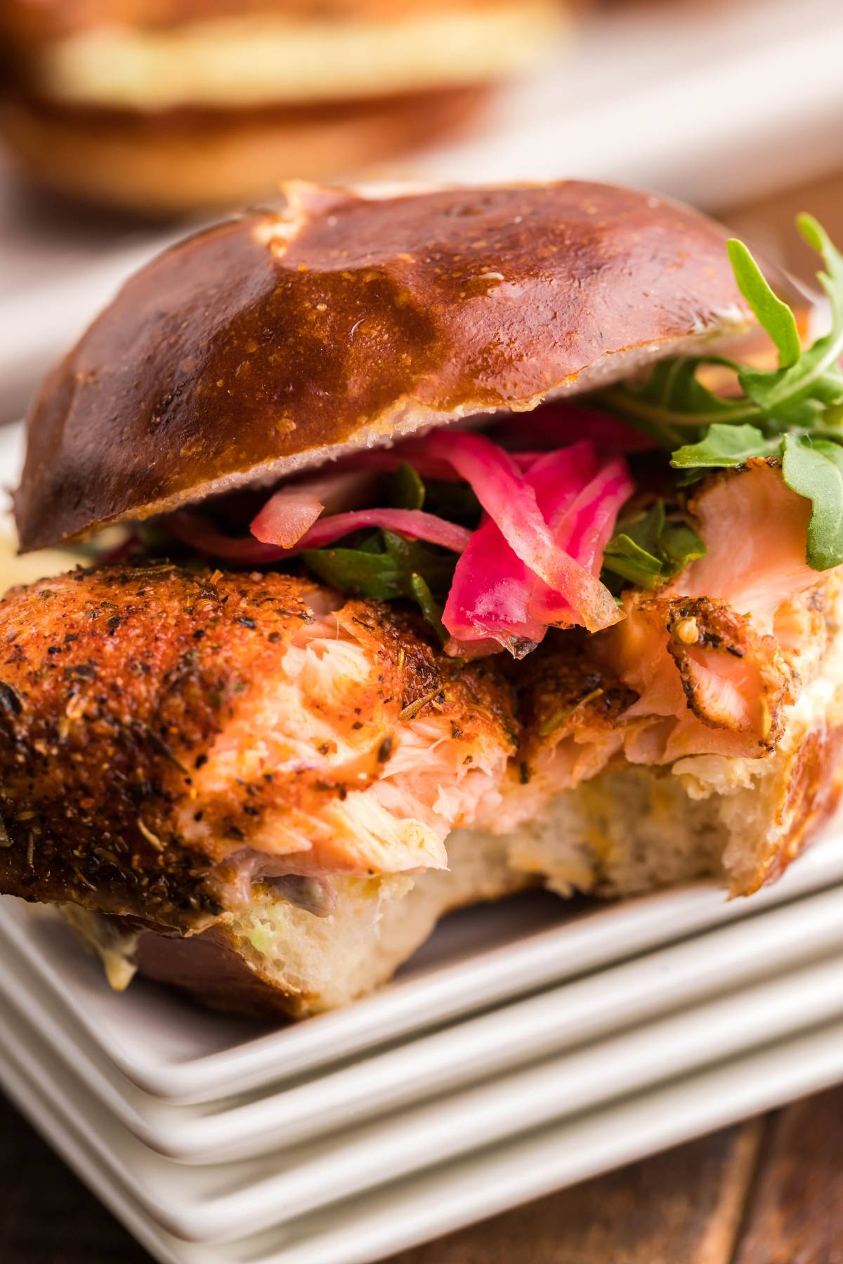 A salmon sandwich with arugula and pickled onions on a pretzel bun, made with our favorite smoked salmon recipe—perfect for Summer BBQ salmon burgers. One tempting bite is already missing!.