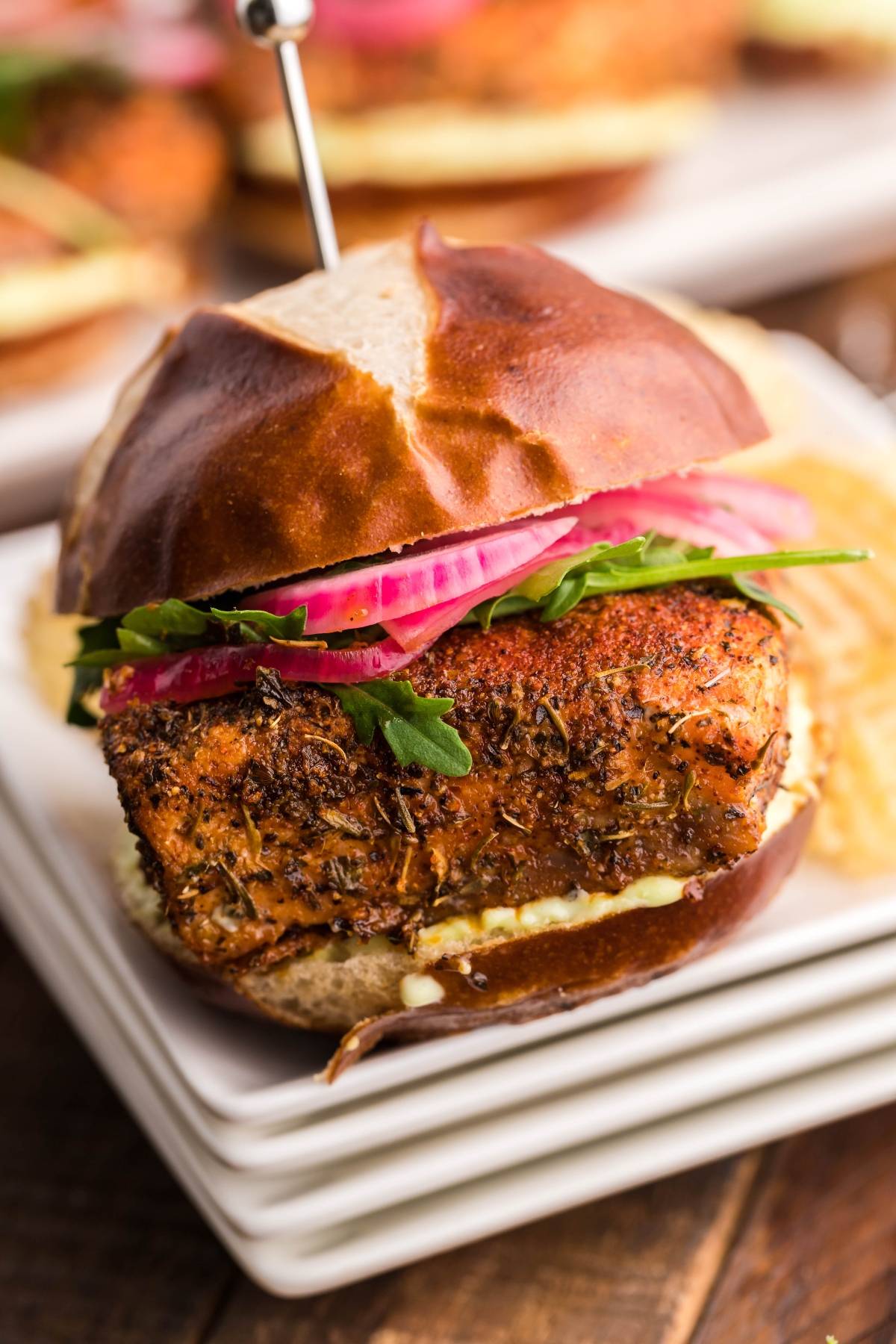 A blackened fish sandwich with arugula, pickled onions, and sauce on a pretzel bun—perfect for fans of Summer BBQ salmon burgers or dishes made in a pellet grill or Traeger.