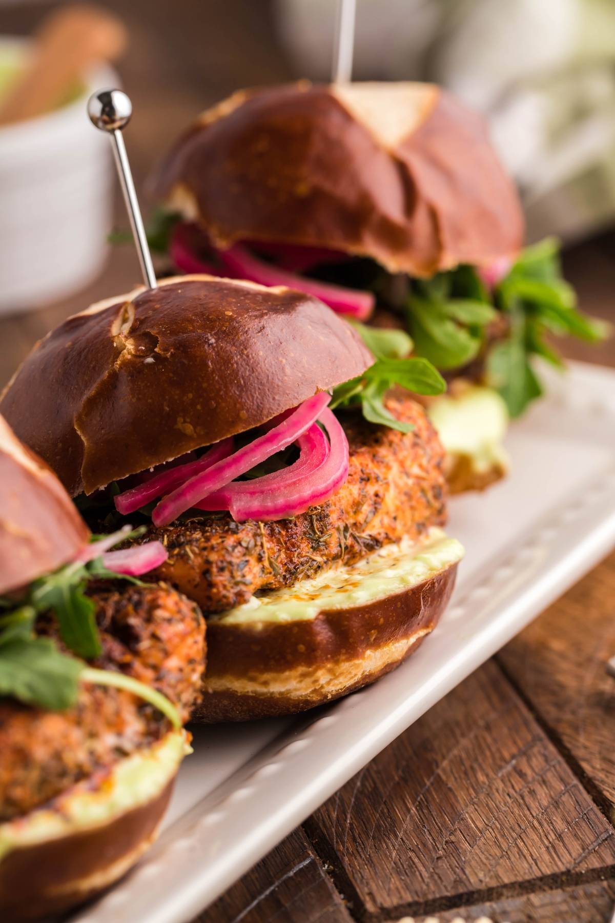 Three veggie burgers with leafy greens and pickled onions on pretzel buns, served on a white rectangular plate—perfect for pairing with Summer BBQ salmon burgers made in a pellet grill or Traeger.