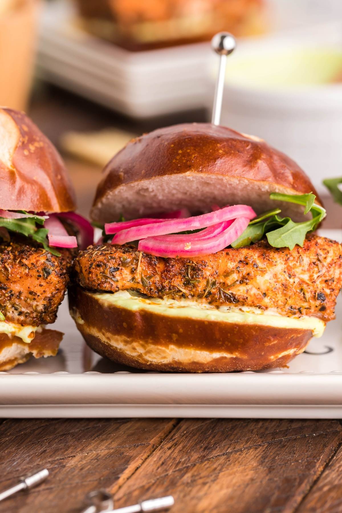 A close-up of a chicken sandwich with lettuce, pickled onions, and a pretzel bun on a plate—perfect alongside Summer BBQ salmon burgers made in a pellet grill or Traeger.