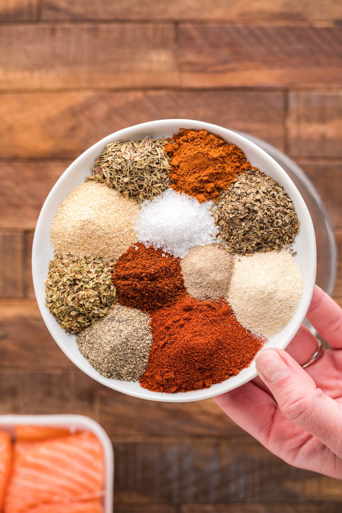 A hand holds a bowl filled with various colorful ground spices and herbs, perfect for seasoning Summer BBQ salmon burgers or smoked salmon sliders, with a wooden surface in the background.
