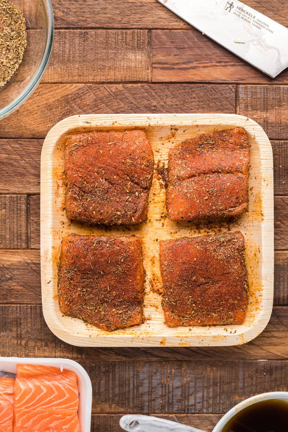 Four raw salmon fillets coated with spices rest on a square plate atop a wooden surface with seasoning nearby—perfect for your next smoked salmon recipe or summer BBQ salmon burgers.
